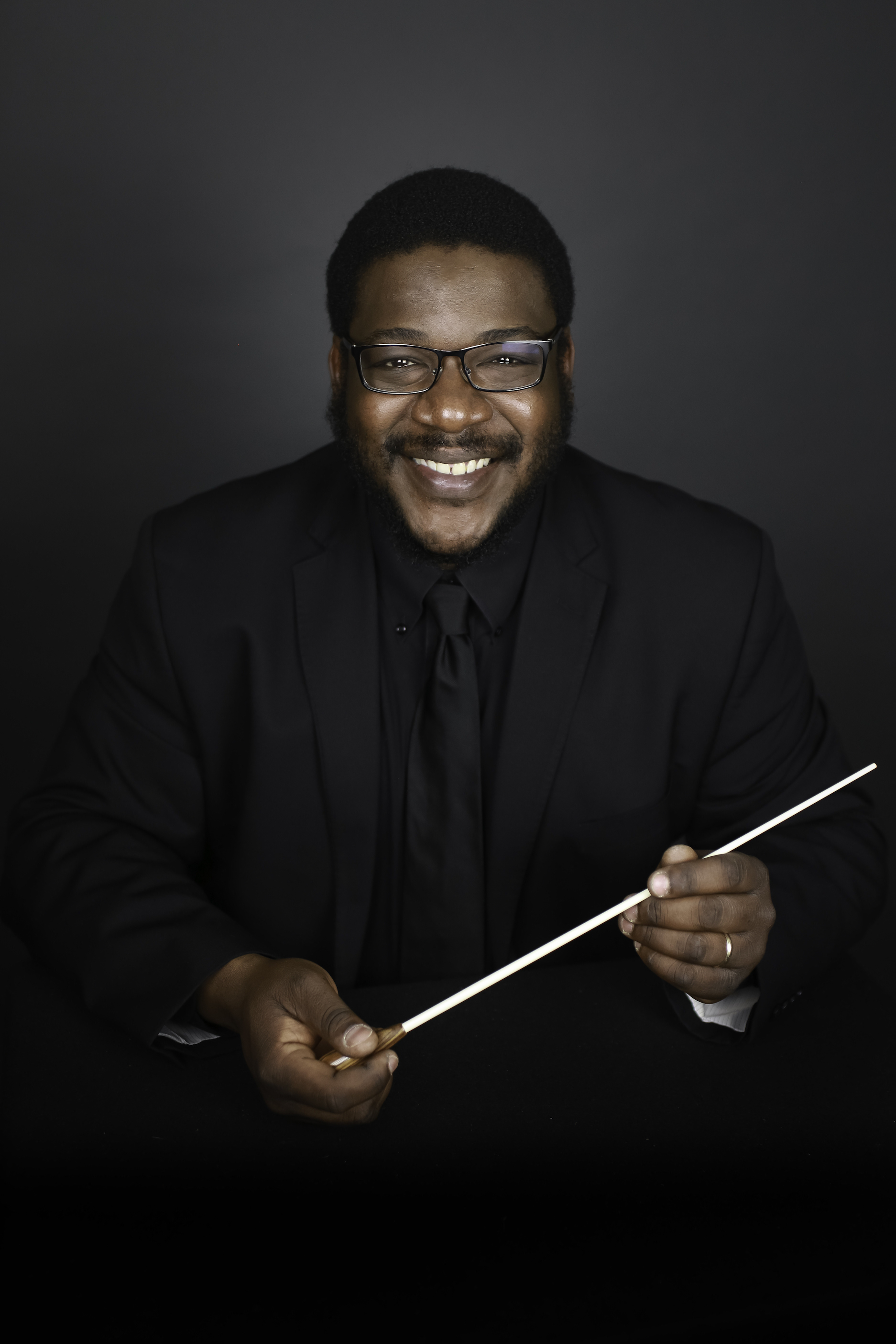 Conductor in concert black holding a white baton, with a dark gray backdrop behind.