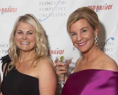 Two people dressed in formal attire, one holding up a martini glass, in front of a brand wall with text: PSO BRAVO!, Princeton Symphony Orchestra