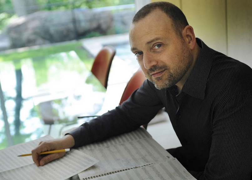 The composer holds a pencil over his score by a sunlit window.