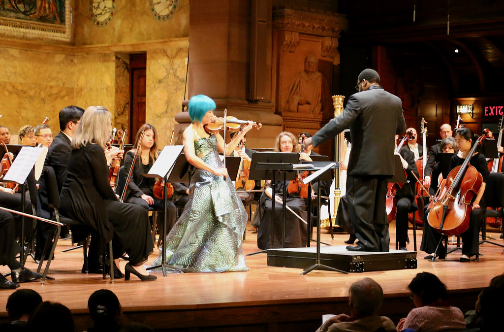 Solo violinist playing onstage beside a conductor on a podium, surrounded by string musicians including violinists, violists, and cellists.