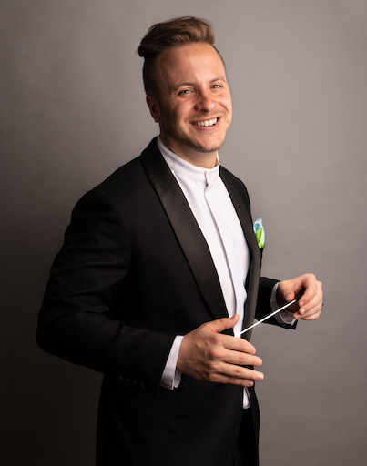 Smiling conductor in a black tuxedo, holding a baton out between both hands.