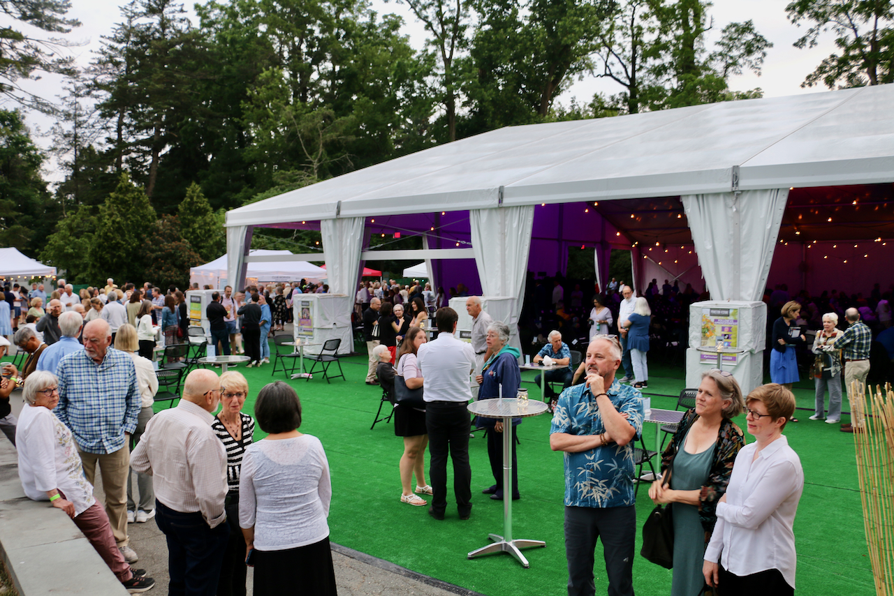 Over 35 people are gathered in small groups outside a large, white, tented structure, with an outdoor gathering area with astroturf flooring