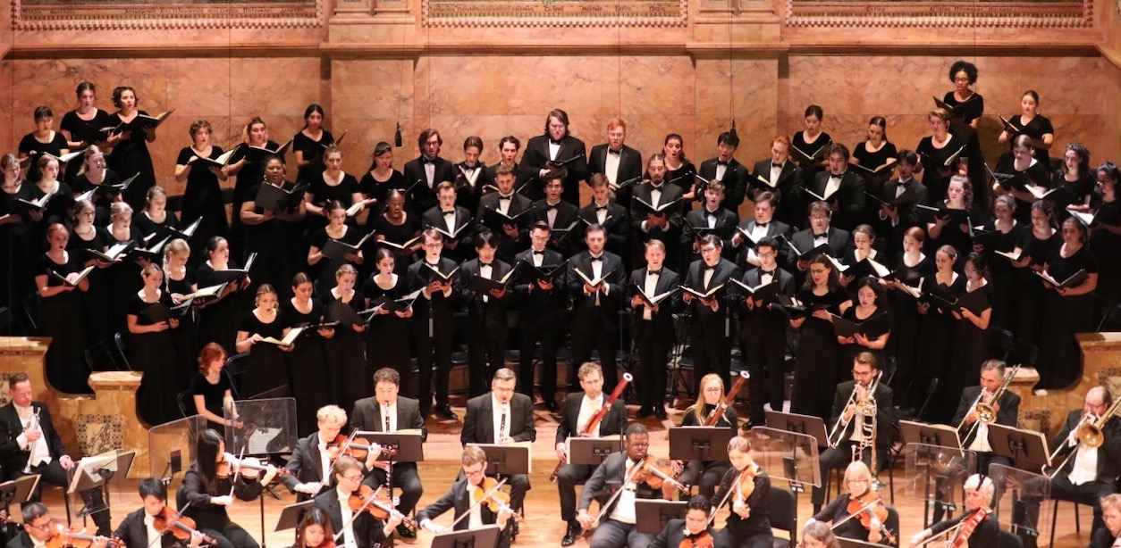 Westminster Symphonic Choir in concert black onstage with music binders, behind rear orchestra during performance.