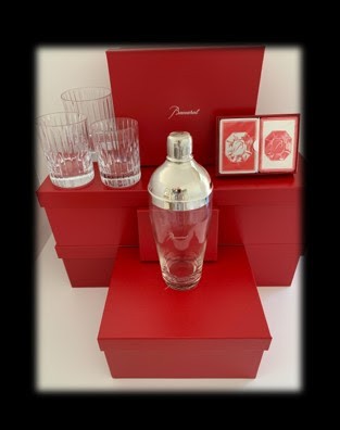 Three sizes of Baccarat crystal tumblers are displayed on top of a red Baccarat box on the left side of the image. On the right side, two boxes of Baccarat playing cards. In the middle, a vintage cocktail shaker.
