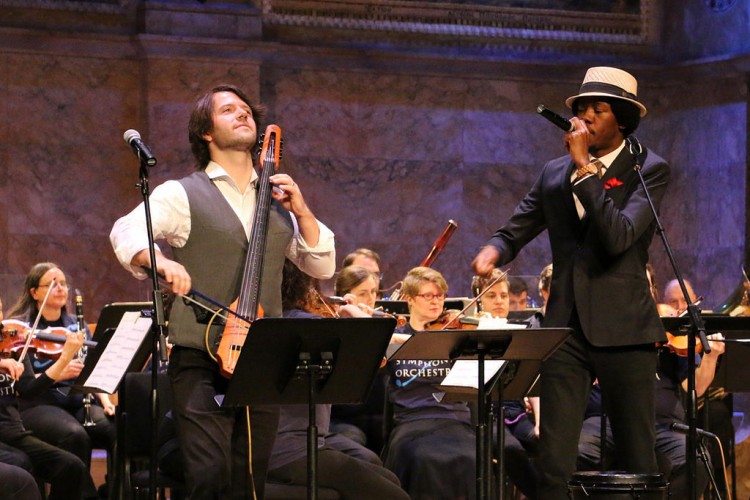 An electric cellist and beat boxer play onstage with orchestra members in blue t-shirts