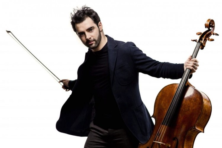 Pablo Ferrández looking directly at the camera with arms spread out. In his left hand he holds a cello. In his right hand, he holds the bow.