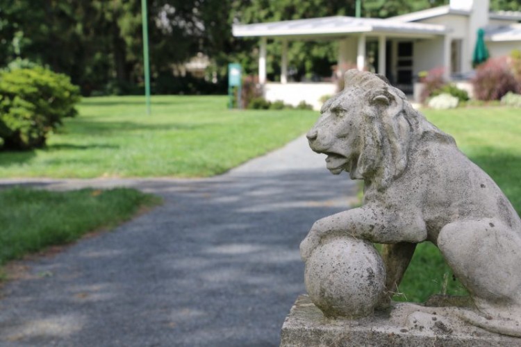 A gravel pathway through a lawn leading up to a building with an open porch. A statue of a lion is next to the pathway.