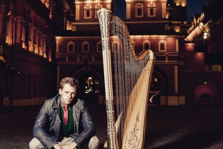Alexander Boldachev poses crouched on the ground next to a harp. It's nighttime. Boldachev and the harp are in front of a lit up building.