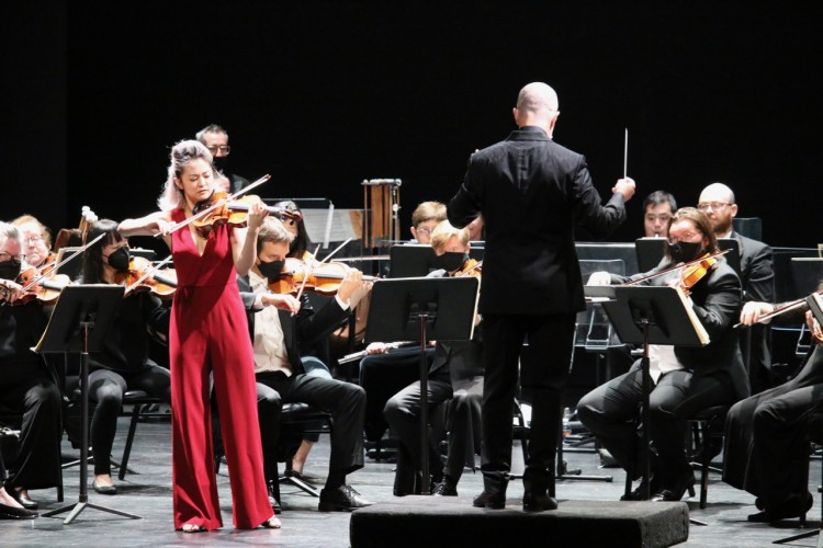 Simone Porter plays the violin on stage. To her right is Rossen Milanov, facing away from the camera and conducting the orchestra. Behind them, the orchestra is currently playing.