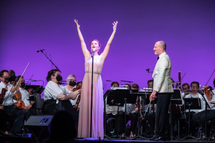 Sierra Boggess stands at a microphone on stage, arms held high above her head as she sings. Behind her are conductor Rossen Milanov and the musicians of the orchestra.