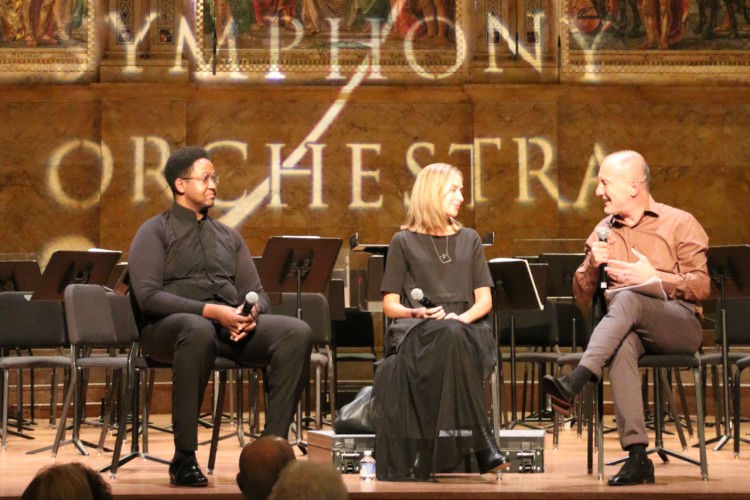 Three people seated onstage with microphones with a projected text on the wall behind: Princeton Symphony Orchestra