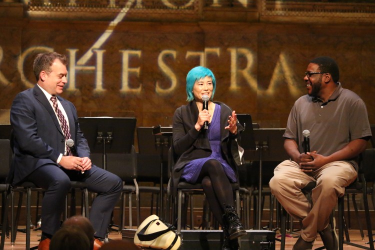 Three people seated onstage with microphones with a projected text on the wall behind: Princeton Symphony Orchestra