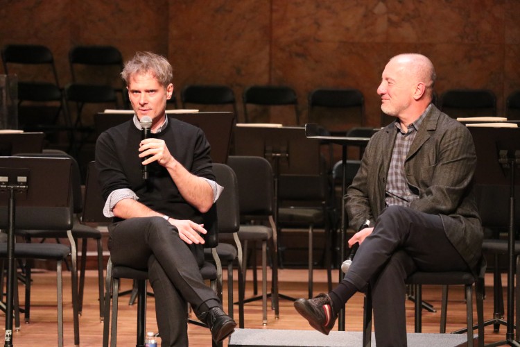 Two people on stage with empty chairs and music stands behind; one is speaking into a microphone while the other looks on.