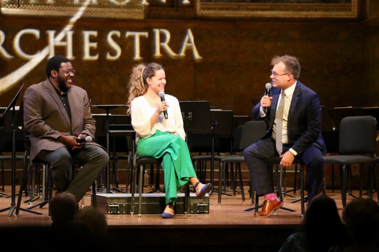 Two men sit on stage with microphones with a younger woman between them as they chat in front of audience.