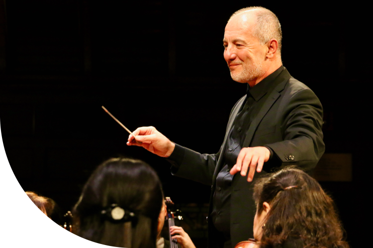 A close up shot of Rossen Milanov conducting. He is holding a baton in his right hand, and his left hand is outstretched toward the violin section. He is wearing a black shirt and jacket. He is smiling slightly. The auditorium behind him is dark. You can see the backs of two musicians' heads in the image's foreground.