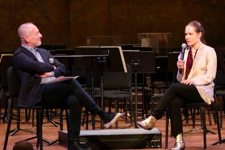 Two people sit on chairs nearly facing each other on a stage holding microphones as they converse.