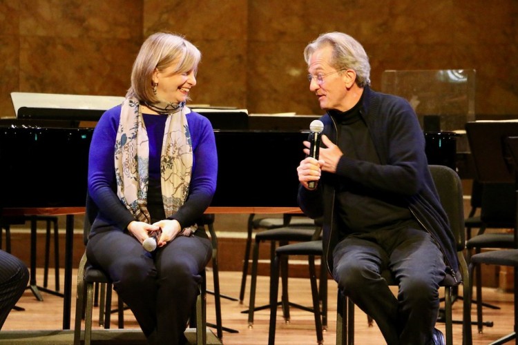 Two people sit on chairs on a stage. A woman holding her hands in her lap smiles to the side as a man with a microphone speaks.