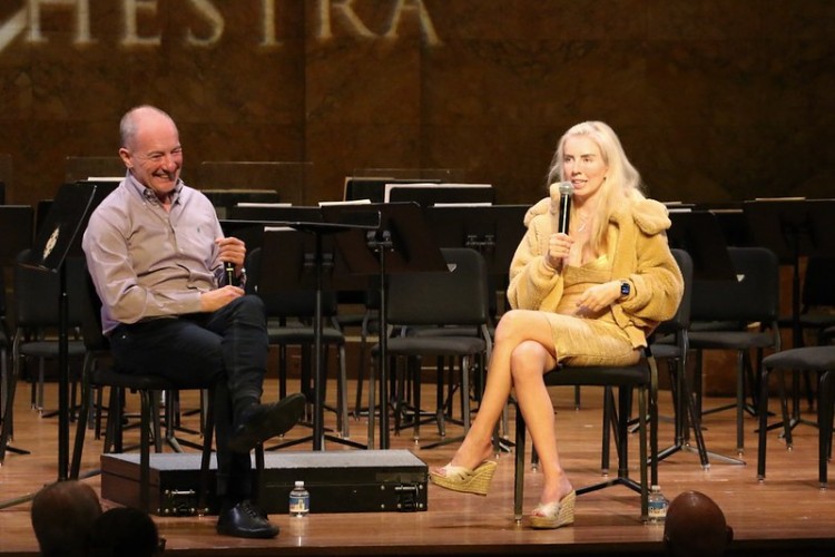 Two people sit on chairs on a stage holding microphones as one laughs and the other speaks. 