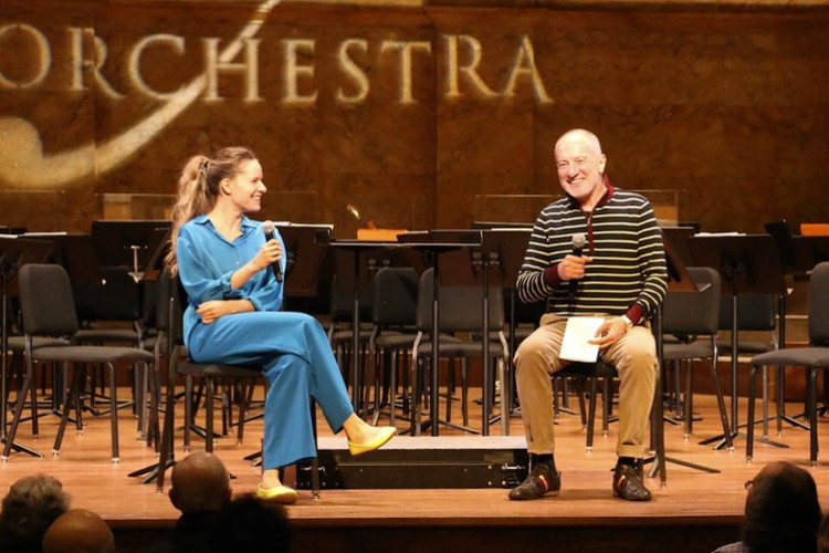 A mature man and a young woman sit on a stage on chairs holding microphones.
