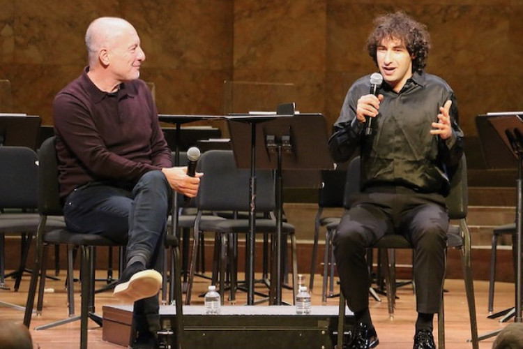 A mature man sits on stage holding a microphone while a young man gestures while speaking into a microphone. 