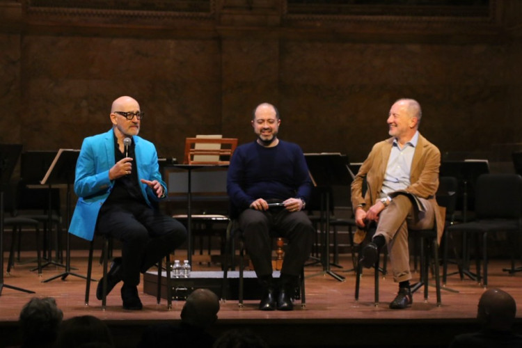 Three men sit on a stage with microphones in front of an audience. 