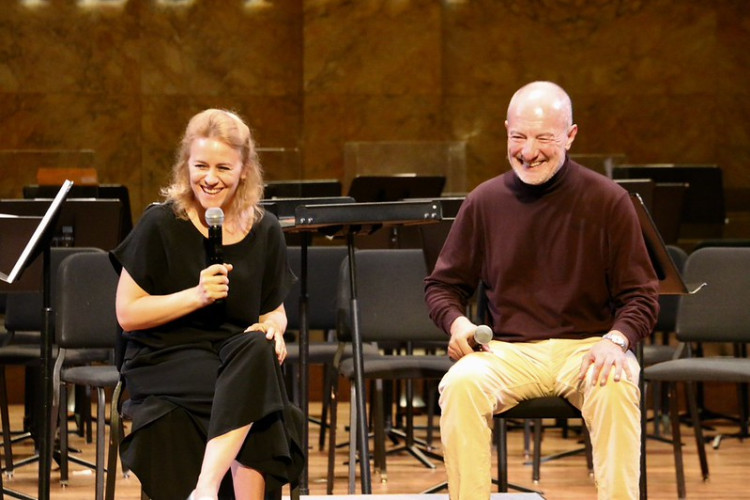 Two people laugh on stage while holding microphones and looking towards the audience. 
