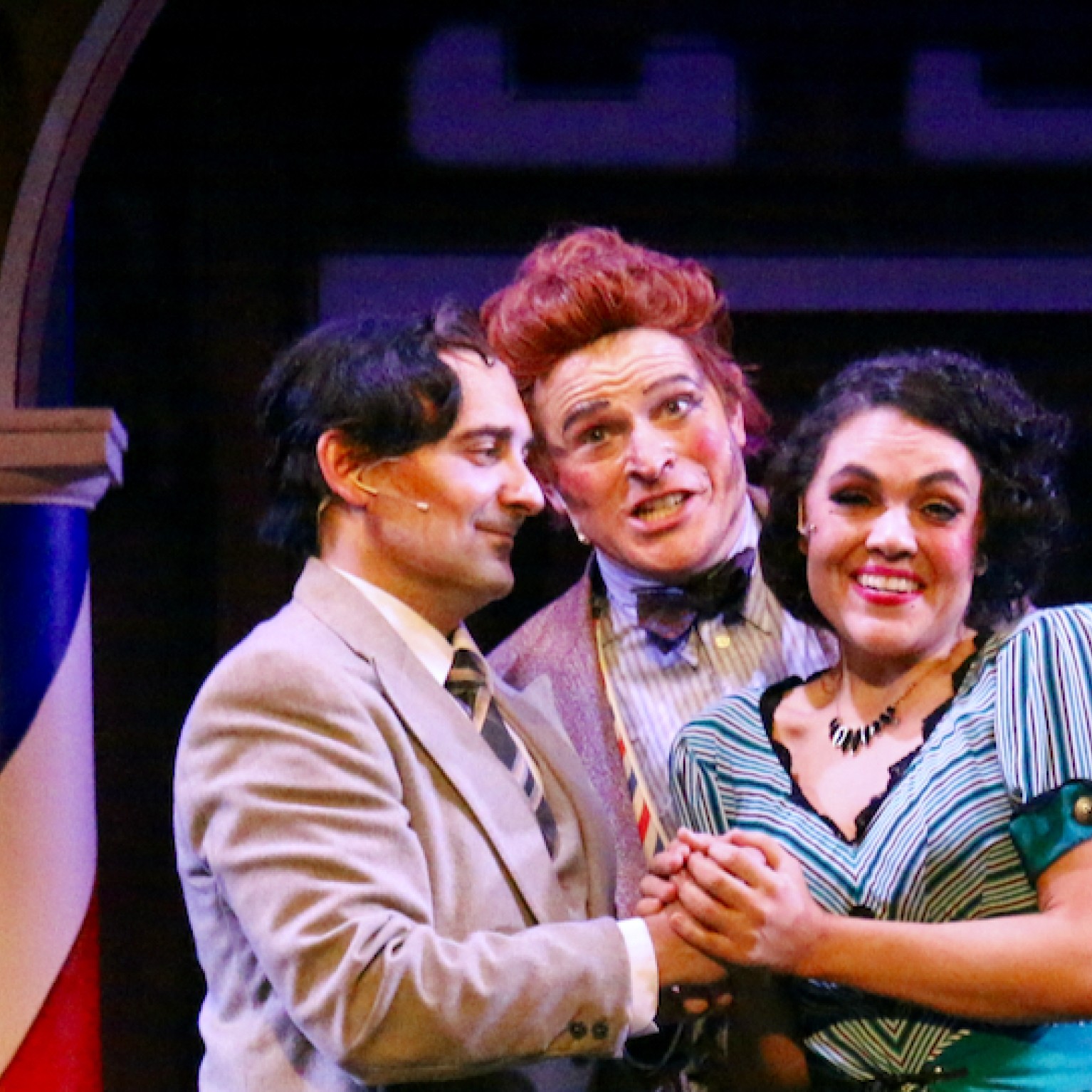 Three costumed opera singers performing onstage between two poles with red, white, and blue barbershop stripe design.