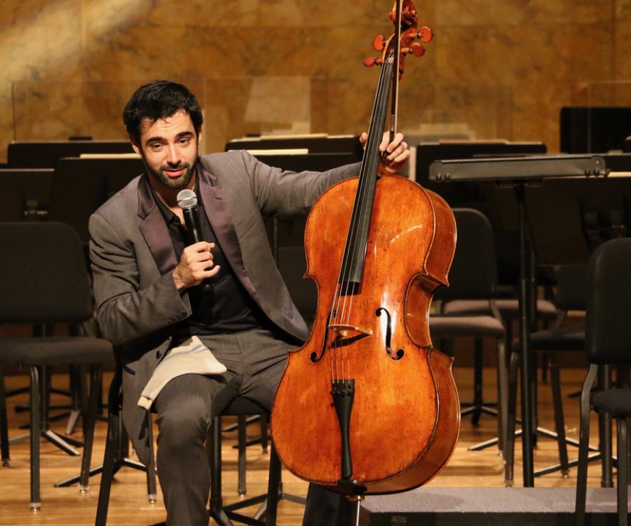 Pablo Ferrández, seated on-stage, holding a cello and speaking into a microphone