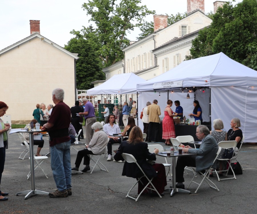 People clustered in groups, sitting and standing, in front of a white concessions canopy with large home-like structures beyond.