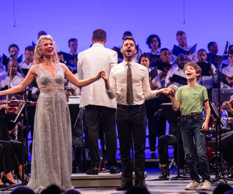 3 singers holding hands up in front of a conductor with orchestra and chorus on stage.