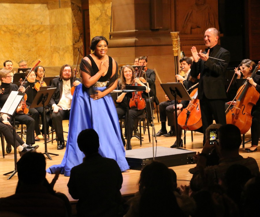 Singer on stage surrounded by a clapping conductor and orchestra members wearing concert black, with silhouetted audience members standing in foreground.