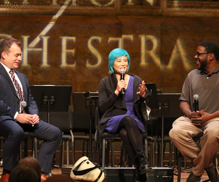 Three smiling people seated on a stage and holding microphones; the center one is talking the others are smiling; projected text behind: Orchestra