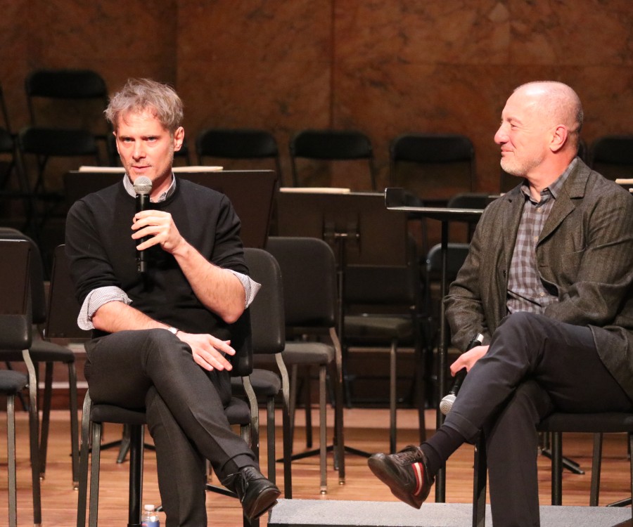 Two people seated onstage with empty chairs and music stands behind; one is speaking into a microphone as the other looks on.