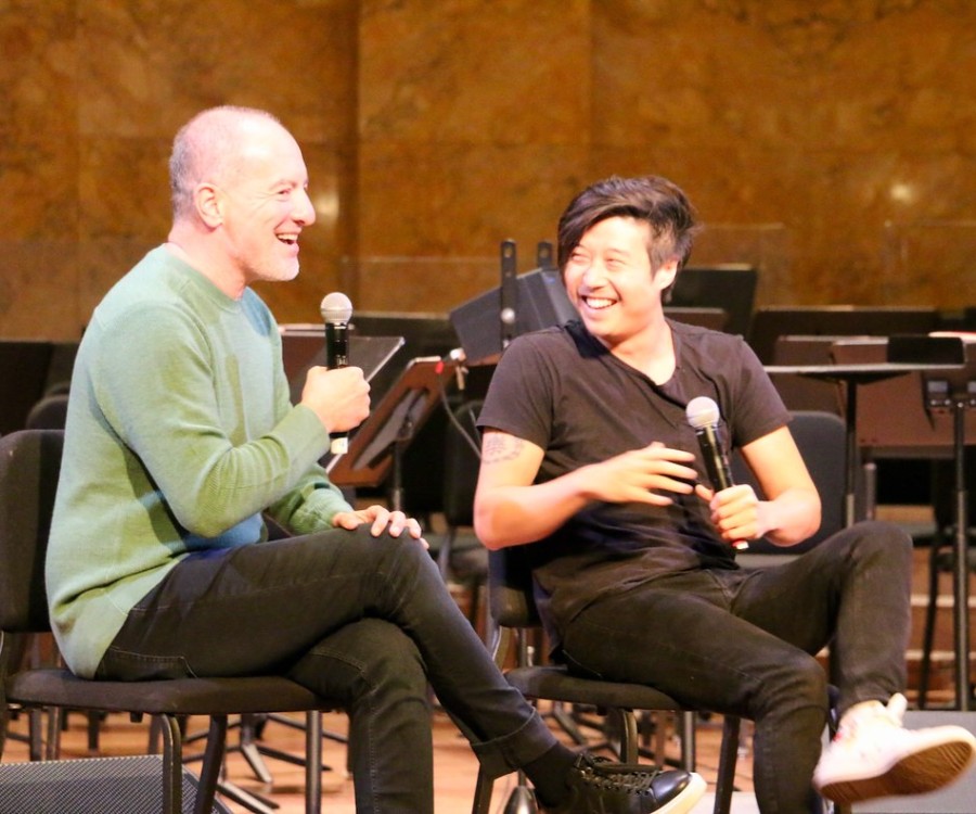 Two people on stage smiling and chatting, both holding microphones. 