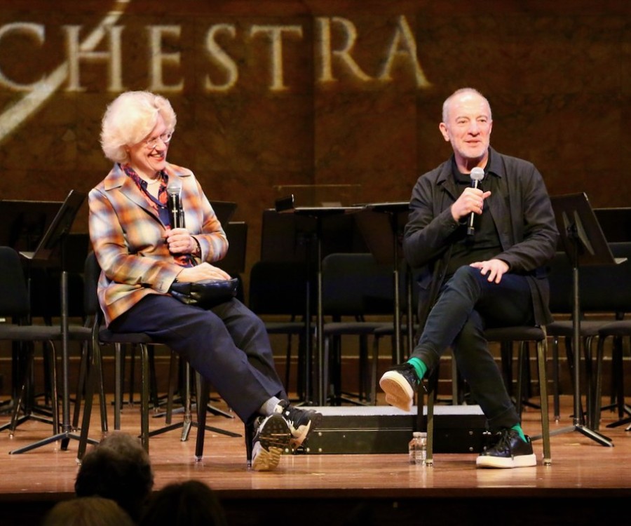 Two people sitting on stage smiling and holding microphones while chatting. 