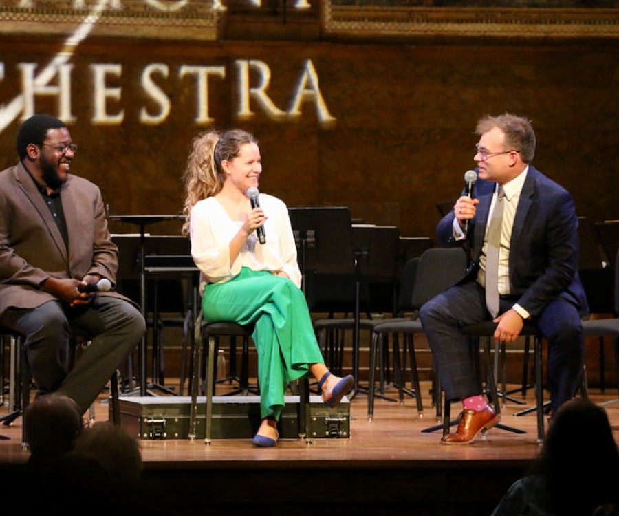 Two men on stage with a younger woman between them holding a microphone as they converse in front of an audience. 