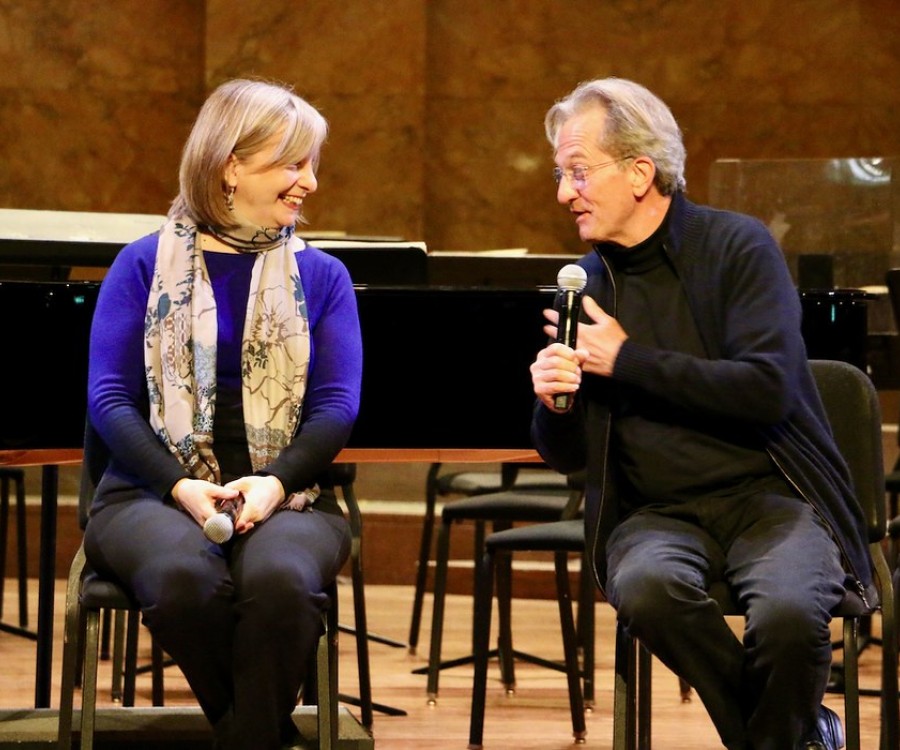 Two people sit on chairs on a stage. A woman holding her hands in her lap smiles to the side as a man with a microphone speaks. 