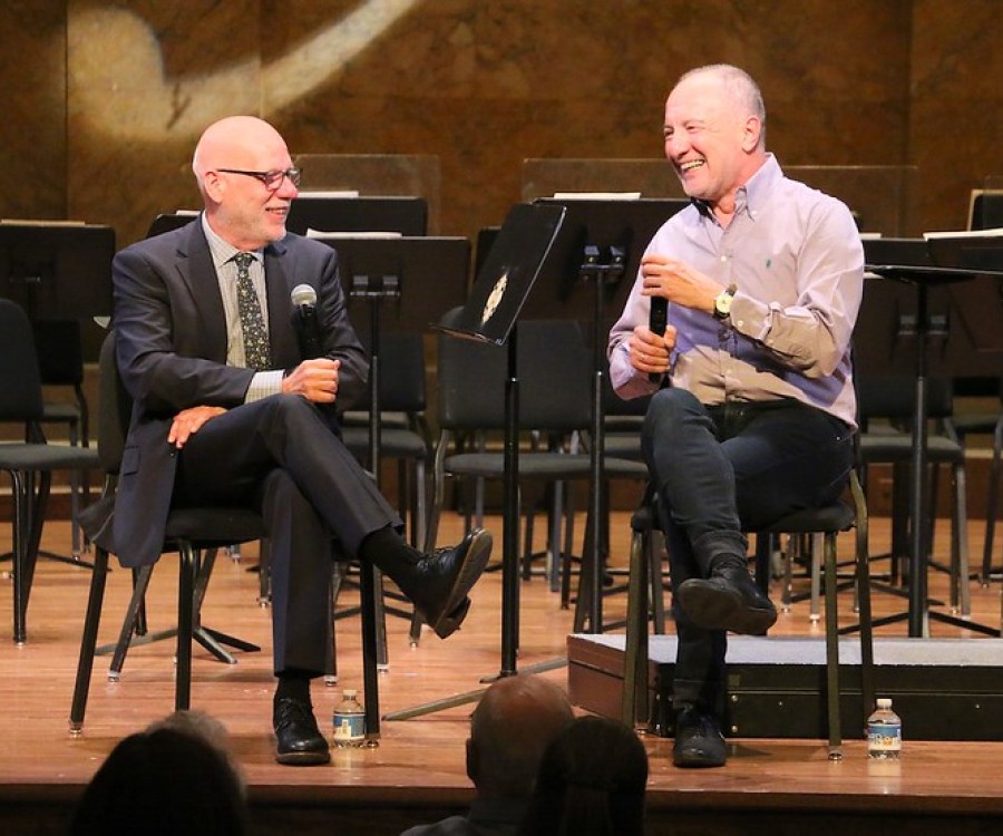 Two men sit on a stage on chairs while holding microphones while talking. 
