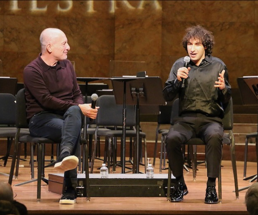 A mature man sits on the stage holding a microphone as he listens to a young man speaking as he gestures.