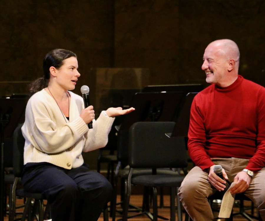 Two people sit on a stage and converse while holding microphones. 