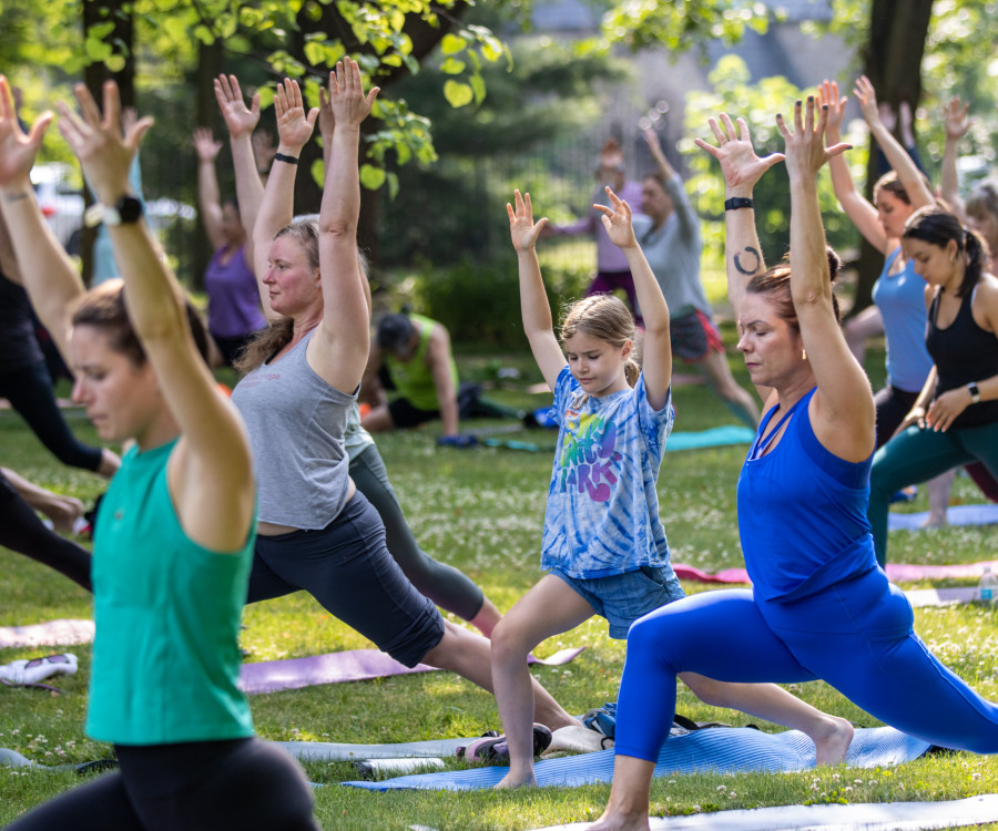 Yoga practitioners working through poses outdoors on a lawn surrounded by tall shade trees.