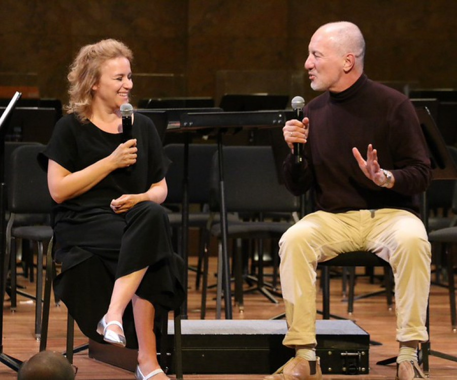 Two people laugh while chatting with each other on a stage with microphones. 