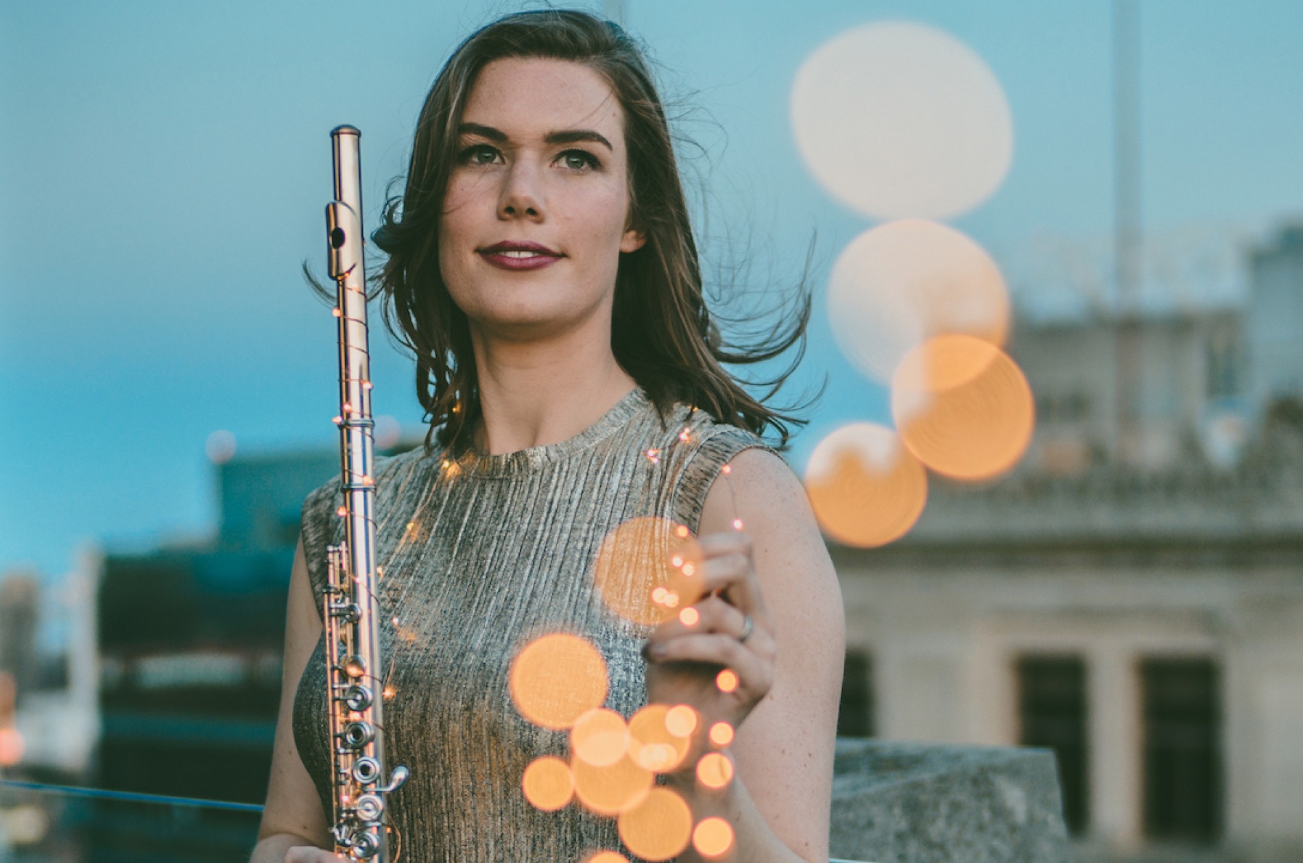 Chelsea Knox holds a flute atop a building, surrounded by orange orbs