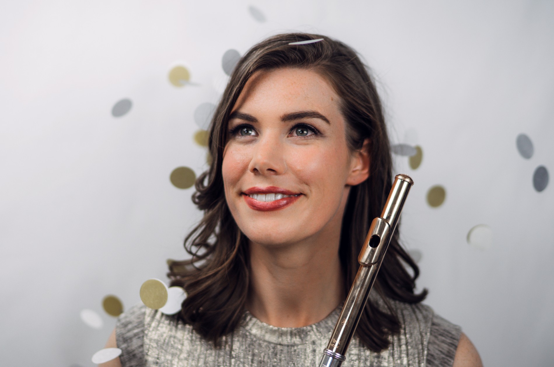 Chelsea Knox stands holding a flute in front of white field with golden discs falling around her