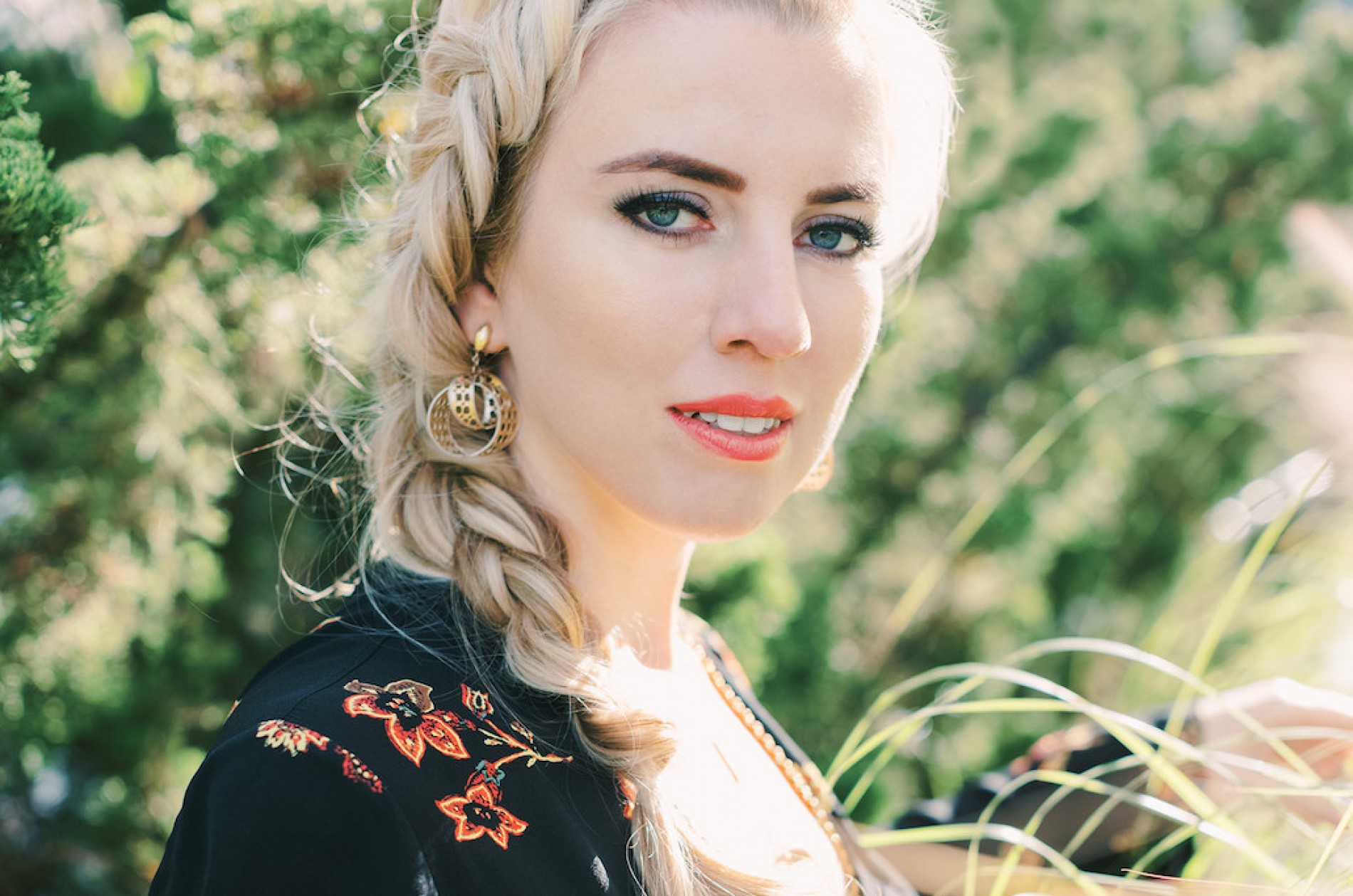 Natasha Paremski stands outside amid flowering branches wearing a black floral print top and a side braid.