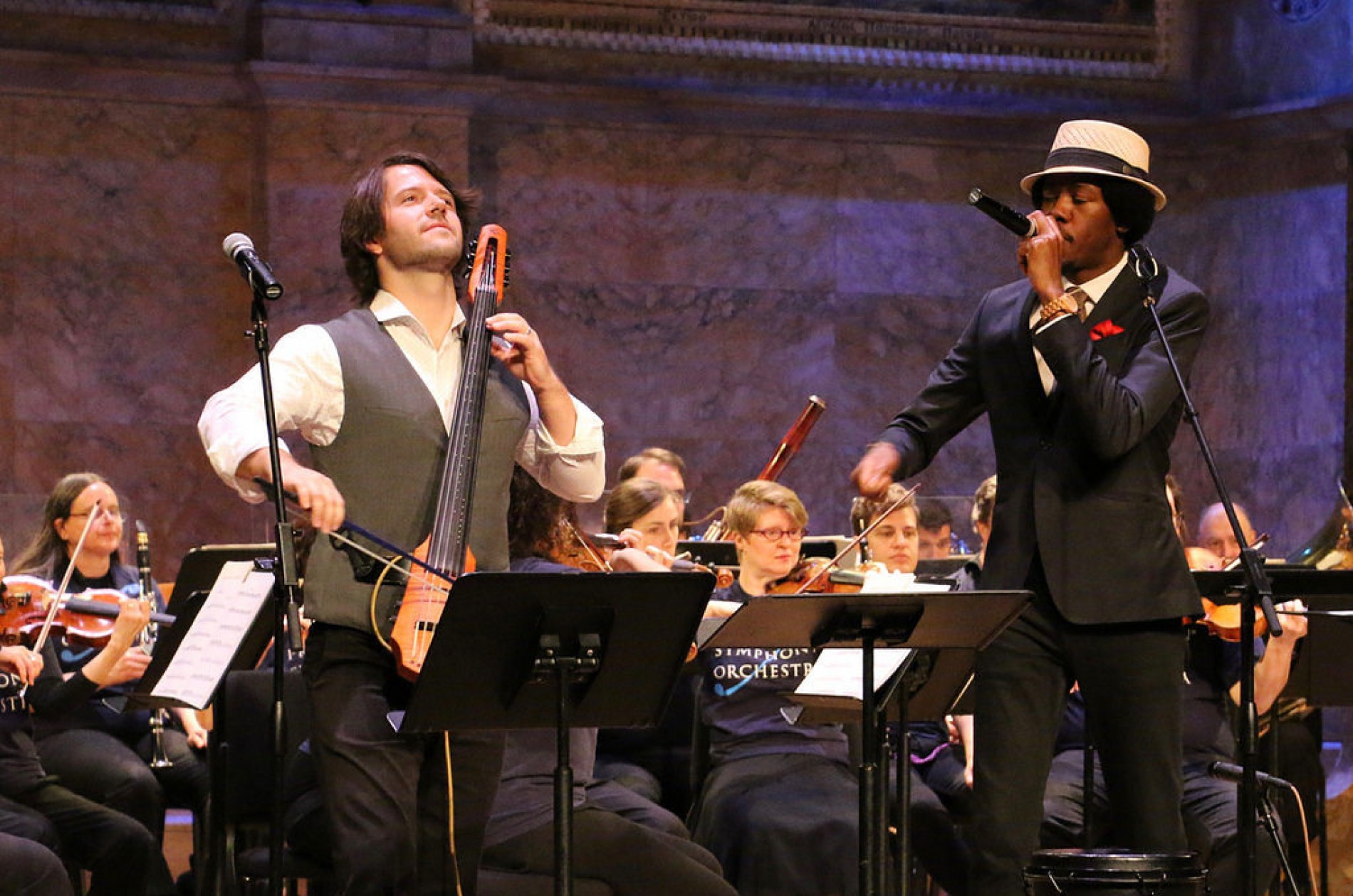 An electric cellist and beat boxer play onstage with orchestra members in blue t-shirts