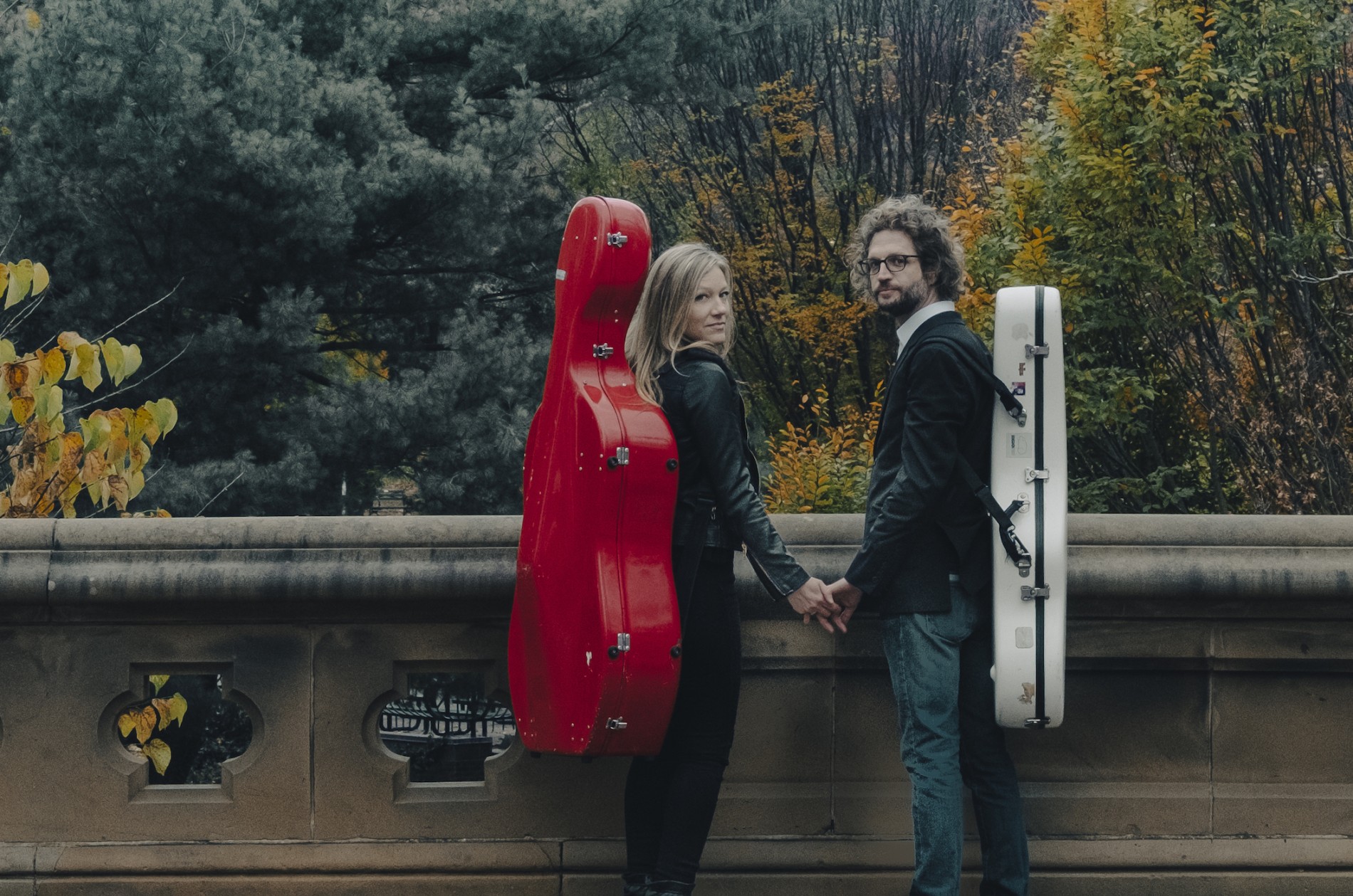 Boyd Meets Girl - a cellist wearing her instrument in a red back-pack carrier and a guitarist wearing his instrument as a back-pack case, holding hands on a bridge in autumn.