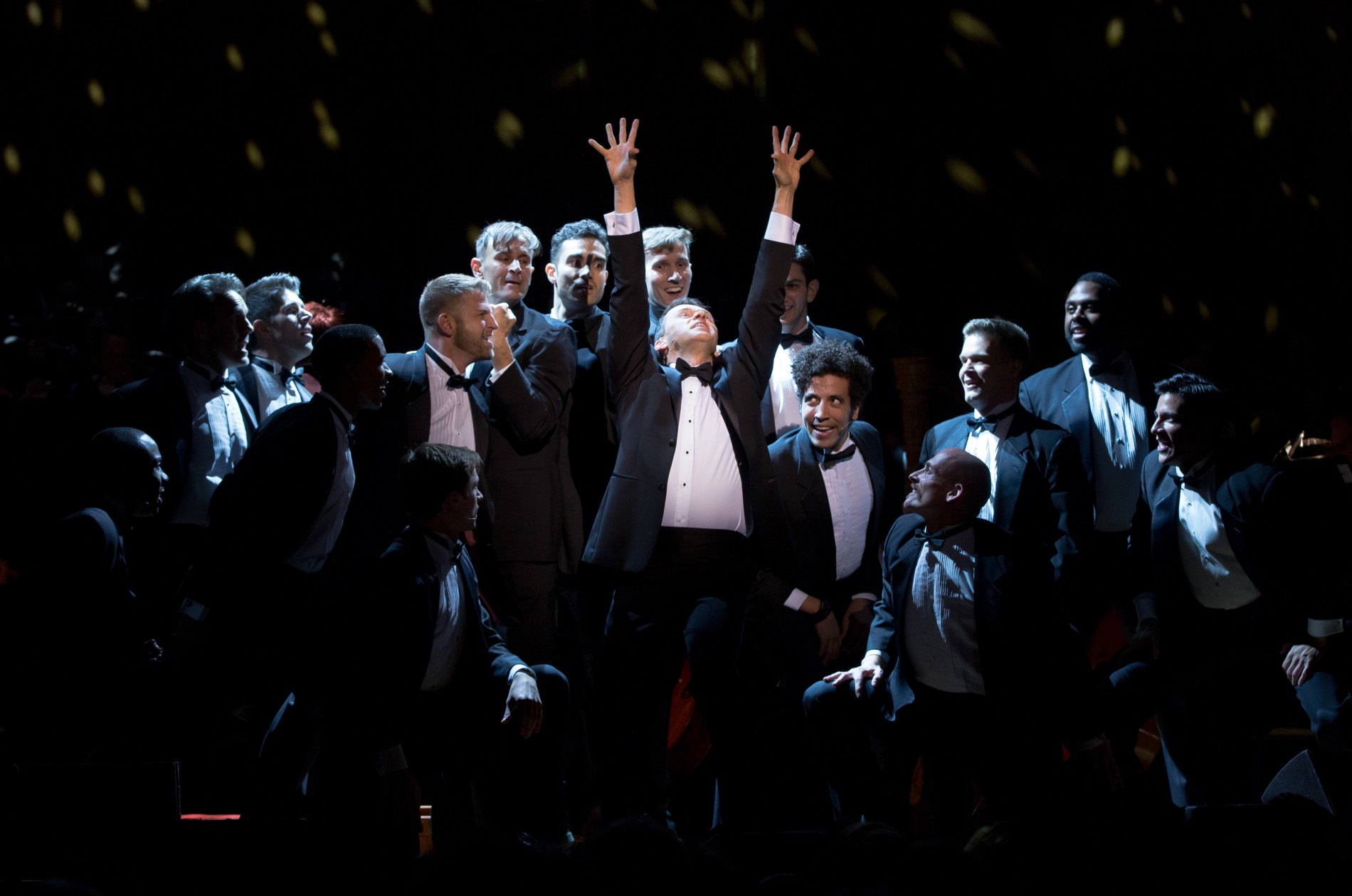 Spotlighted singer in black tuxedo reaching and looking up, surrounded by other artists in tuxedos