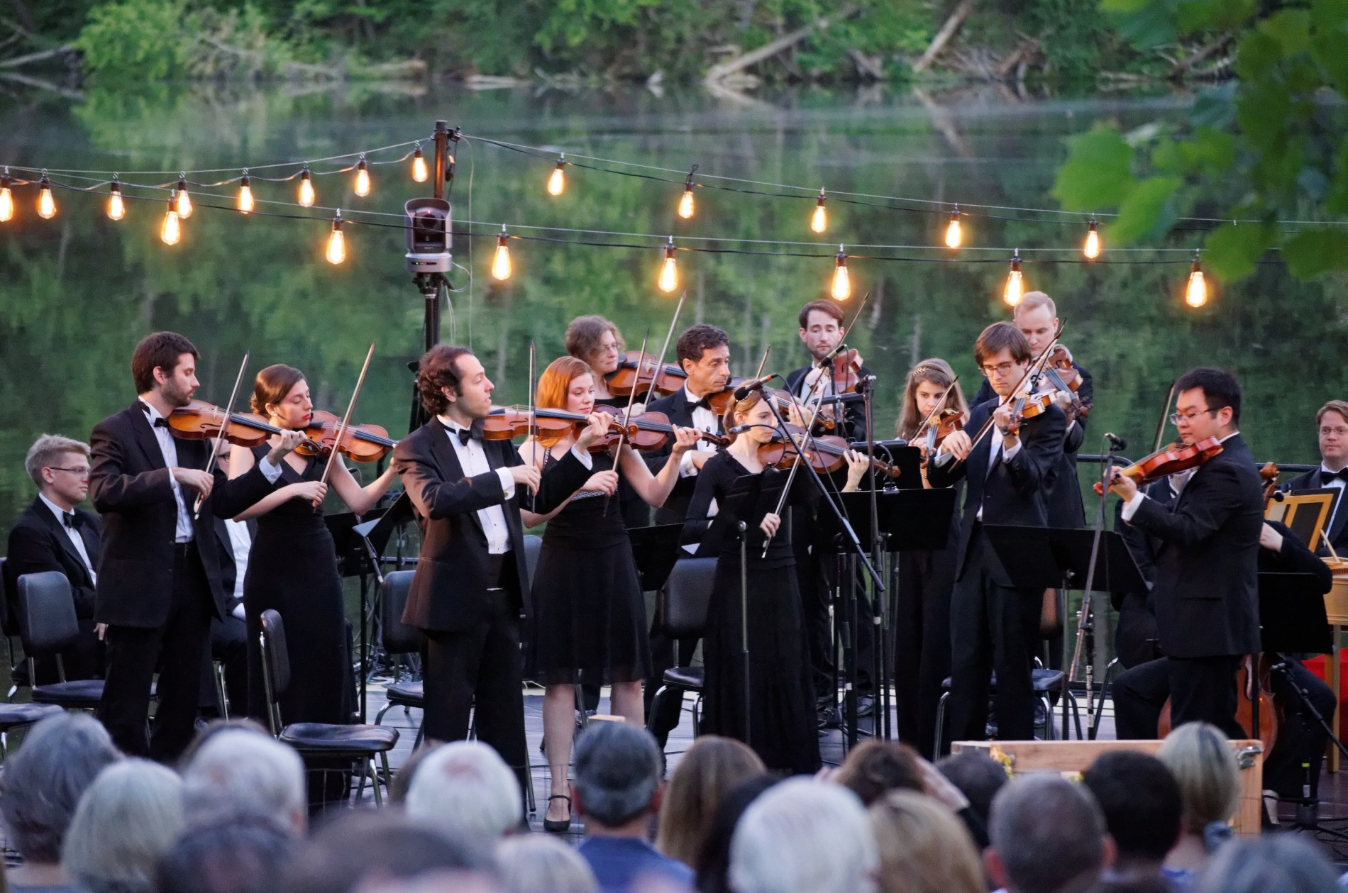 Musicians wearing concert black, standing, playing Baroque instruments in an outdoor setting.