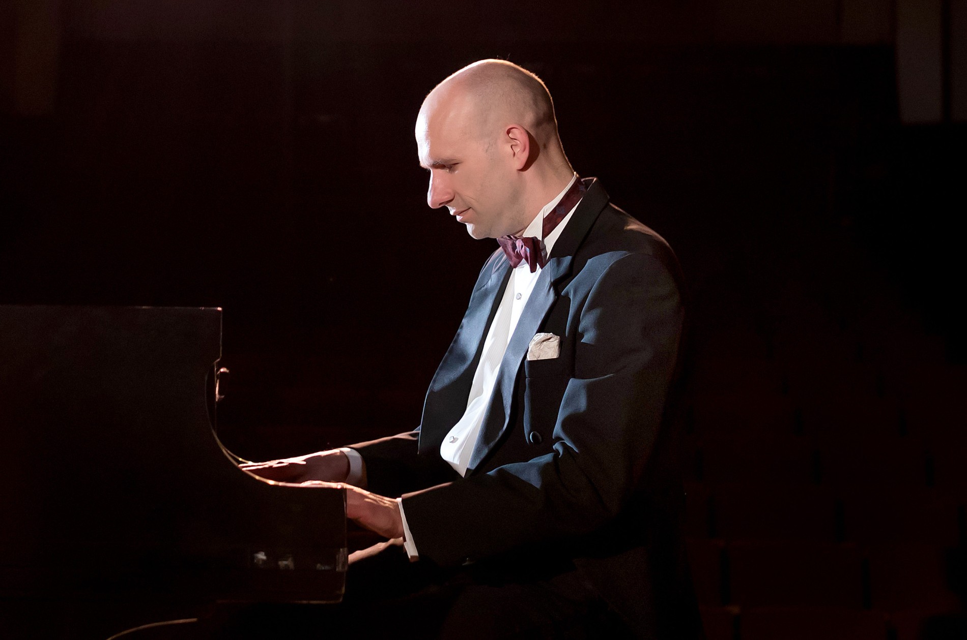 Christopher Taylor, playing the piano in a black tuxedo against a dark background.